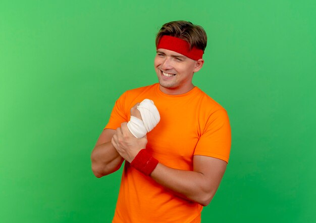 Smiling young handsome sporty man wearing headband and wristbands holding his injured wrist wrapped with bandage isolated on green wall