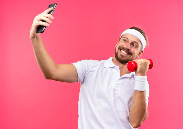 Free photo smiling young handsome sporty man wearing headband and wristbands holding mobile phone and dumbbell and looking at phone isolated on pink space