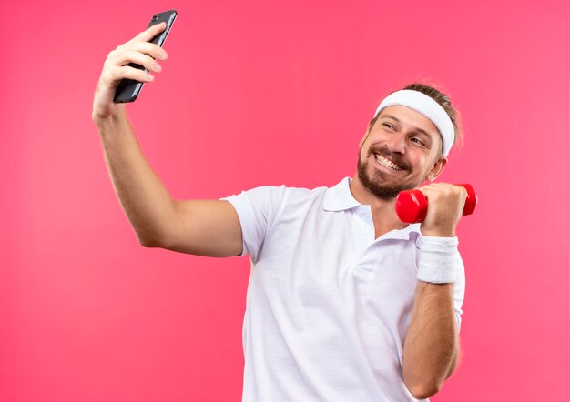 Smiling young handsome sporty man wearing headband and wristbands holding mobile phone and dumbbell and looking at phone isolated on pink space