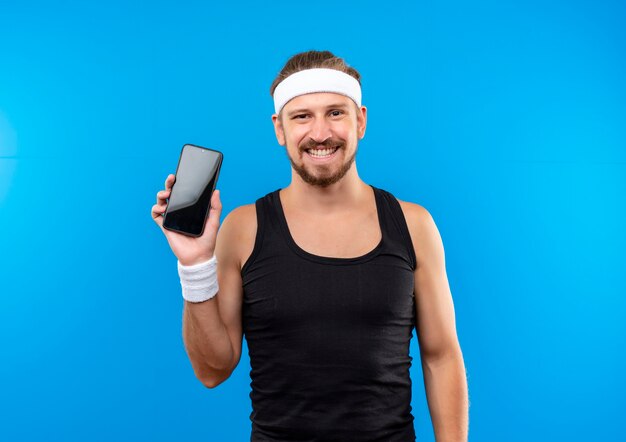 Smiling young handsome sporty man wearing headband and wristbands holding mobile phone isolated on blue space