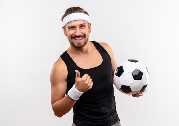 Smiling young handsome sporty man wearing headband and wristbands holding soccer ball and showing thumb up isolated on white space