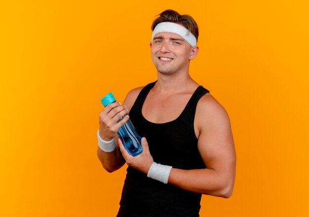 Smiling young handsome sporty man wearing headband and wristbands holding water bottle isolated on orange wall