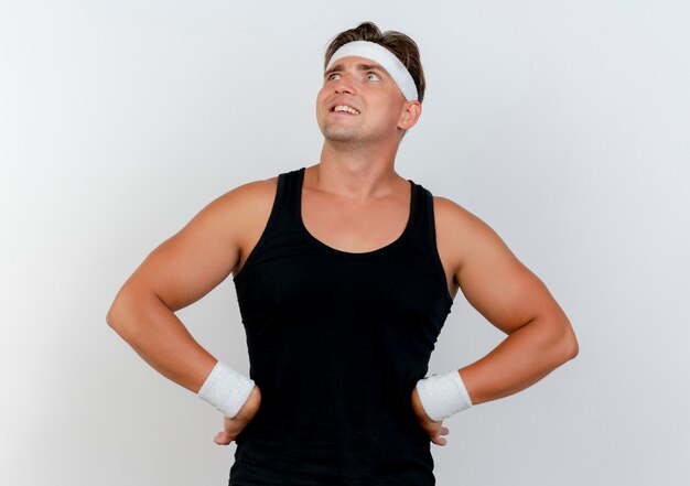Smiling young handsome sporty man wearing headband and wristbands looking up with hands on waist isolated on white wall