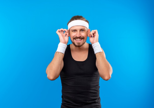Smiling young handsome sporty man wearing headband and wristbands making big ears isolated on blue space 