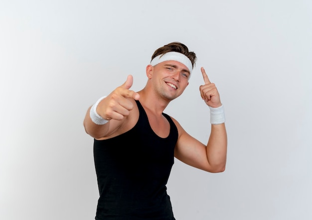 Smiling young handsome sporty man wearing headband and wristbands pointing up and at front isolated on white wall