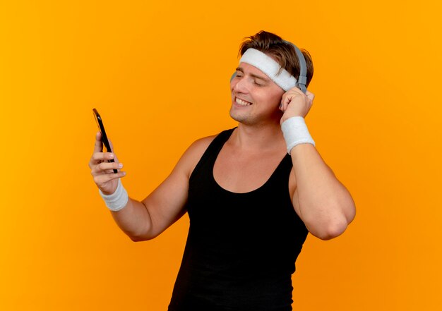 Smiling young handsome sporty man wearing headband and wristbands putting hand on headphones holding and looking at mobile phone isolated on orange wall