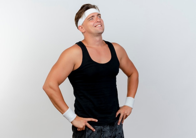 Smiling young handsome sporty man wearing headband and wristbands putting hands on waist looking straight isolated on white wall