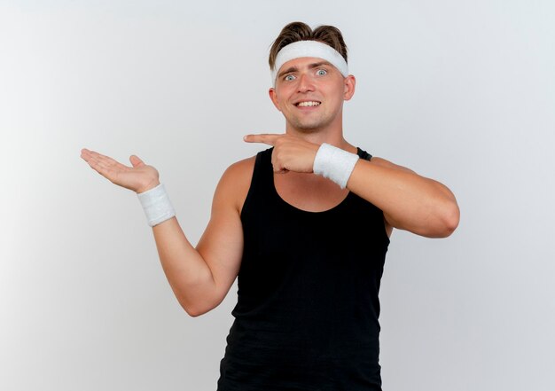 Smiling young handsome sporty man wearing headband and wristbands showing empty hand and pointing at it isolated on white wall