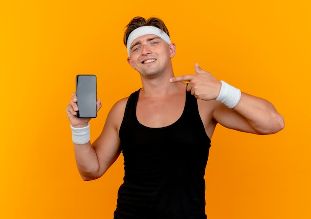 Smiling young handsome sporty man wearing headband and wristbands showing and pointing at mobile phone isolated on orange wall