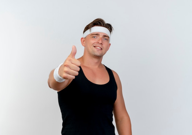 Smiling young handsome sporty man wearing headband and wristbands showing thumb up at front isolated on white wall