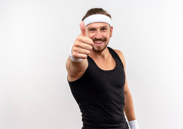 Smiling young handsome sporty man wearing headband and wristbands showing thumb up  isolated on white space 