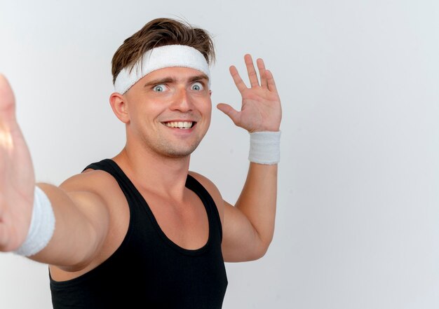 Smiling young handsome sporty man wearing headband and wristbands stretching out hand at front and keeping another one on air isolated on white wall