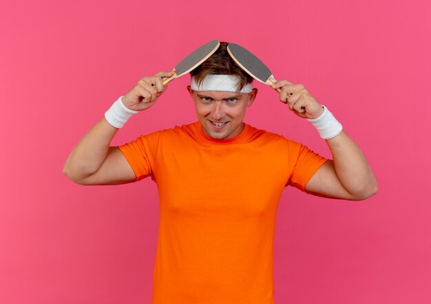 Smiling young handsome sporty man wearing headband and wristbands touching head with ping pong rackets isolated on pink wall