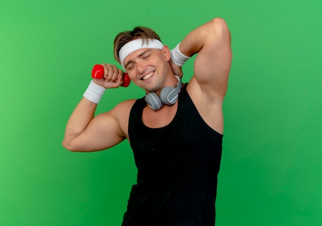 Free photo smiling young handsome sporty man wearing headband and wristbands with headphones on neck holding dumbbell and putting hand on neck isolated on green wall