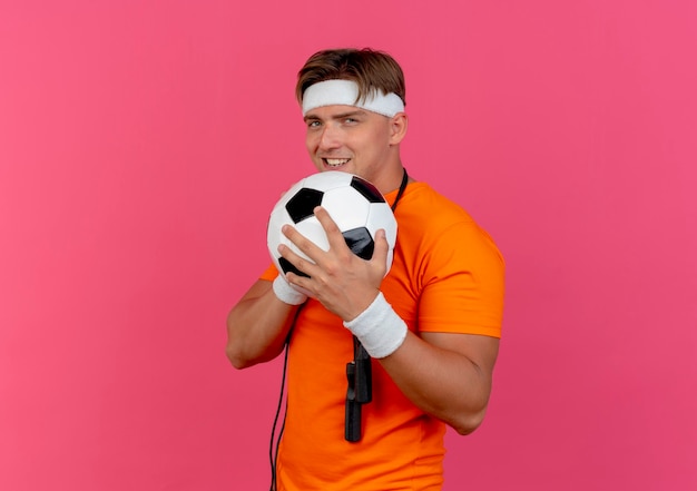 Smiling young handsome sporty man wearing headband and wristbands with jump rope around neck holding soccer ball isolated on pink wall