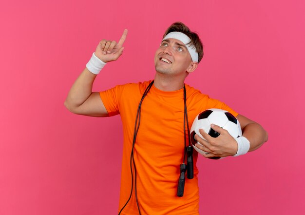 Smiling young handsome sporty man wearing headband and wristbands with jump rope around neck holding soccer ball looking and pointing up isolated on pink wall