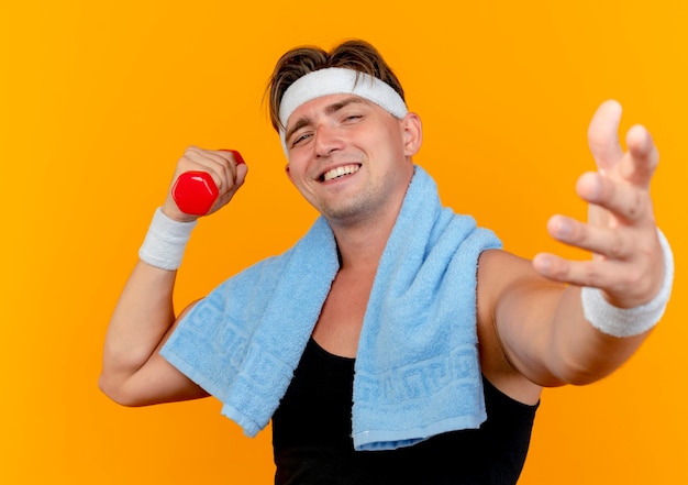 Smiling young handsome sporty man wearing headband and wristbands with towel around neck holding dumbbell stretching out hand at front isolated on orange wall