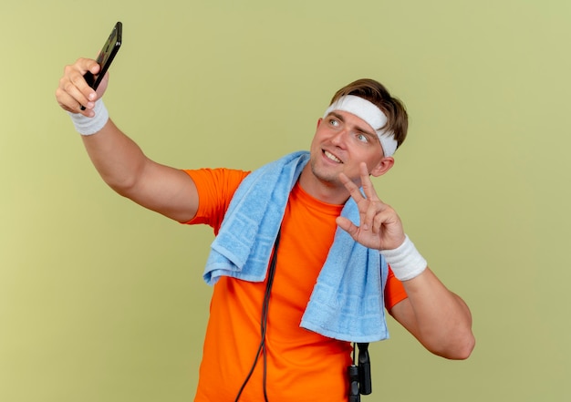 Free photo smiling young handsome sporty man wearing headband and wristbands with towel and jump rope around neck doing peace sign taking selfie isolated on olive green wall