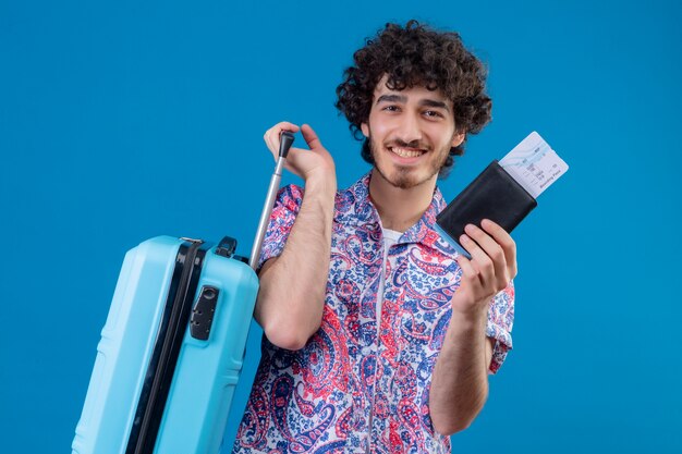 격리 된 파란색 벽에 가방, 비행기 티켓 및 지갑을 들고 웃는 젊은 잘 생긴 여행자 남자