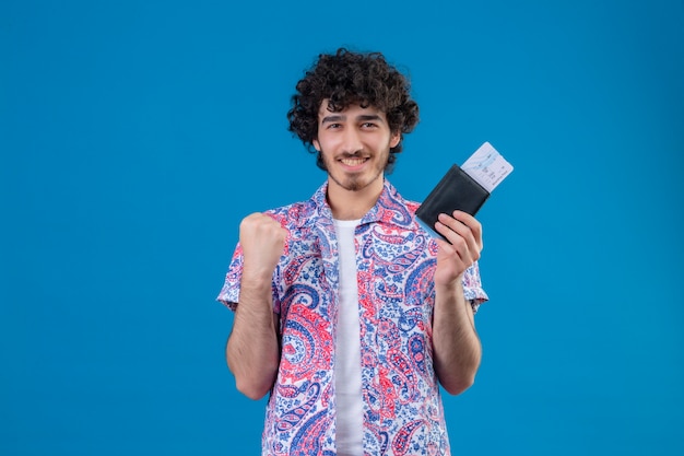 Smiling young handsome traveler man holding wallet and airplane tickets with raised fist on isolated blue wall with copy space