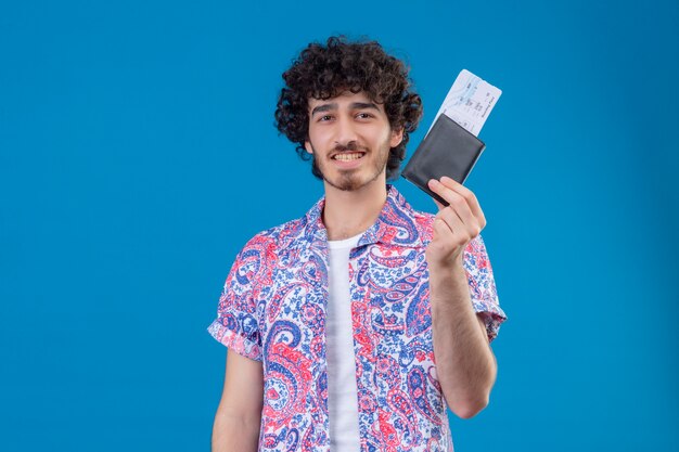 Smiling young handsome traveler man showing wallet and airplane tickets on isolated blue wall with copy space