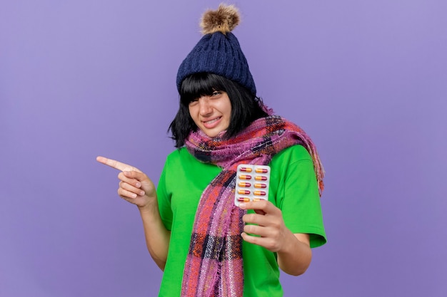 Smiling young ill caucasian girl wearing winter hat and scarf holding pack of capsules looking at camera pointing at side isolated on purple background with copy space