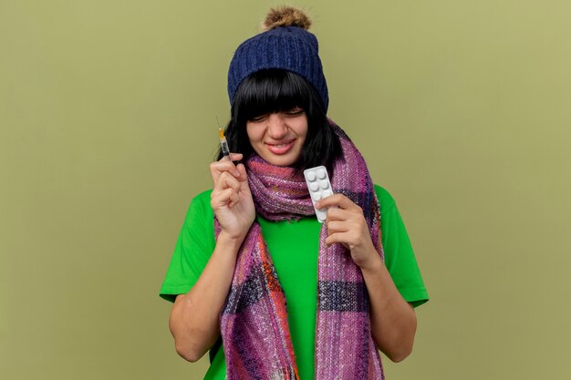 Улыбающаяся молодая больная кавказская девушка в зимней шапке и шарфе держит шприц и упаковку медицинских таблеток с закрытыми глазами, изолированными на оливково-зеленом фоне с копией пространства