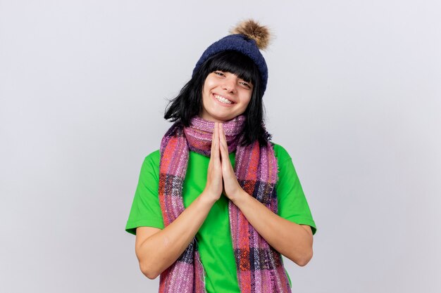 Smiling young ill caucasian girl wearing winter hat and scarf keeping hands together looking at camera isolated on white background with copy space