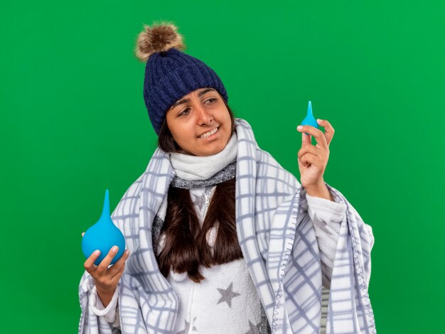 Smiling young ill girl wearing winter hat with scarf holding and looking at enemas isolated on green background