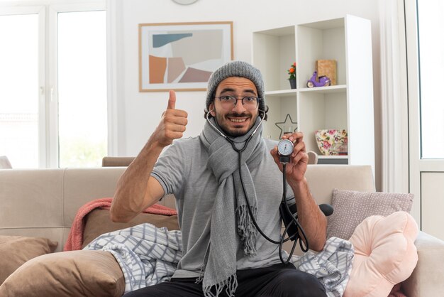 smiling young ill man in optical glasses with scarf around his neck wearing winter hat measuring pressure with sphygmomanometer and thumbing up sitting on couch at living room
