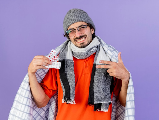 Smiling young ill man wearing glasses winter hat and scarf wrapped in plaid holding and pointing at packs of medical pills looking at front isolated on purple wall