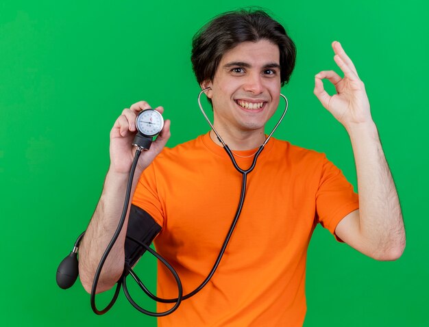 Улыбающийся молодой больной человек со стетоскопом, держащий сфигмоманометр, показывающий жест окей, изолированный на зеленом фоне