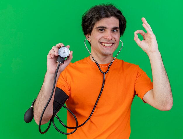 Smiling young ill man wearing stethoscope holding sphygmomanometer showing okey gesture isolated on green background