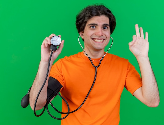 Smiling young ill man wearing stethoscope holding sphygmomanometer showing okey gesture isolated on green background