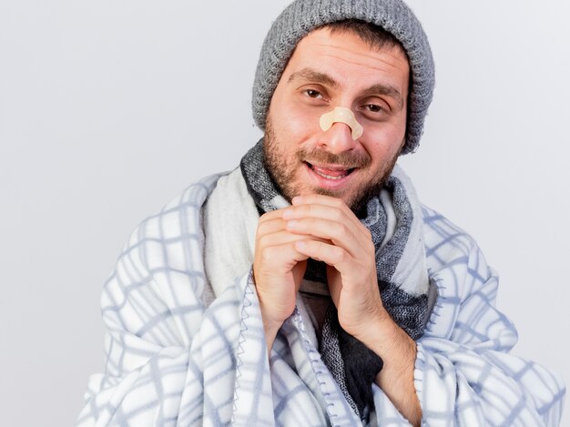 Free photo smiling young ill man wearing winter hat and scarf wrapped in plaid with plaster on nose holding hands on chin isolated on white