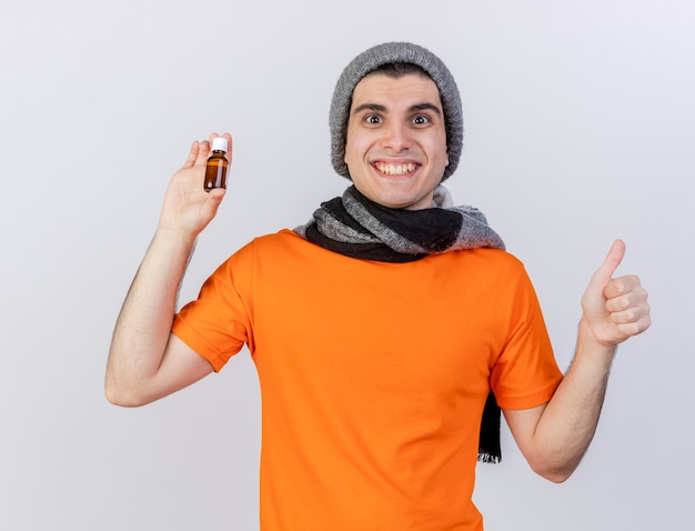 Smiling young ill man wearing winter hat with scarf holding medicine in glass bottle showing thumb up