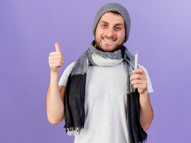 Smiling young ill man wearing winter hat with scarf holding thermometer showing thumb up isolated on purple