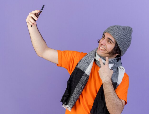 Smiling young ill man wearing winter hat with scarf take a selfie showing peace gestureisolated on purple background
