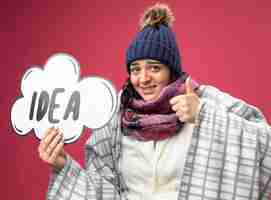 Free photo smiling young ill woman wearing robe winter hat and scarf wrapped in plaid holding idea bubble looking at front showing thumb up isolated on pink wall