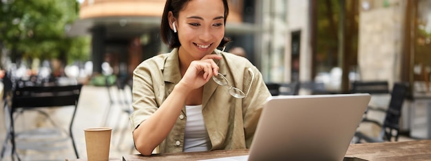 Smiling young korean woman looks at her laptop screen with pleased face works remotely from outdoors