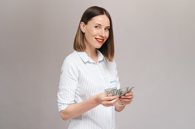 Free photo smiling young lady in striped shirt holding money and looking to them over gray wall looking to front