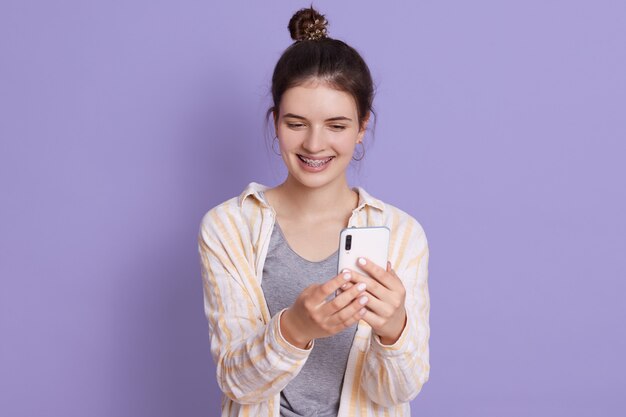 Smiling young lady with hair bun holding modern smart phone in hands and making selfie