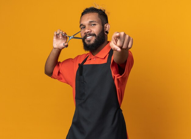 Smiling young male barber wearing uniform holding scissors looking and pointing at front isolated on orange wall with copy space