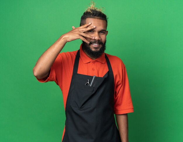 Smiling young male barber wearing uniform keeping hand on face looking at front through fingers isolated on green wall with copy space