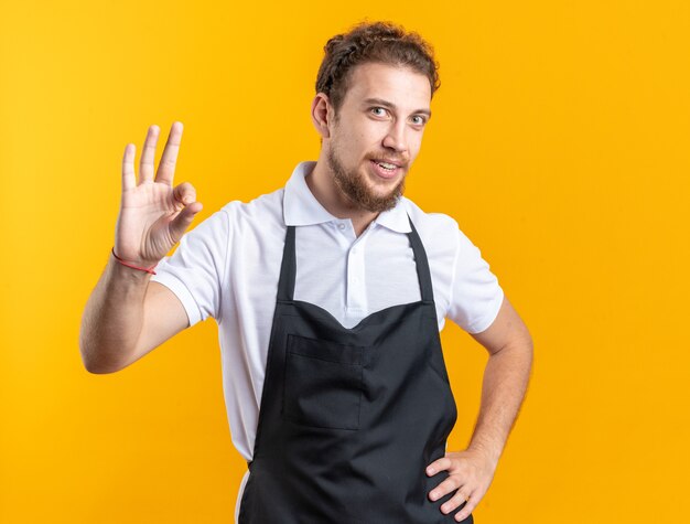 Smiling young male barber wearing uniform showing okay gesture putting hand on hip isolated on yellow background