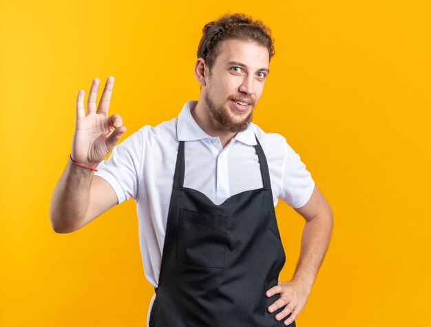 Улыбающийся молодой мужчина-парикмахер в униформе показывает нормальный жест, положив руку на бедро, изолированные на желтом фоне