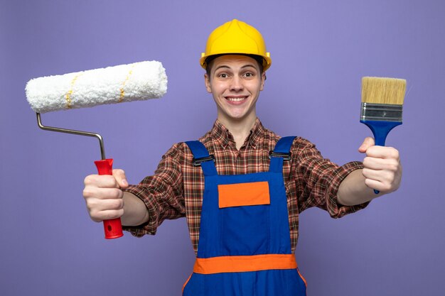 Улыбающийся молодой мужчина-строитель в униформе держит кисть с валиком