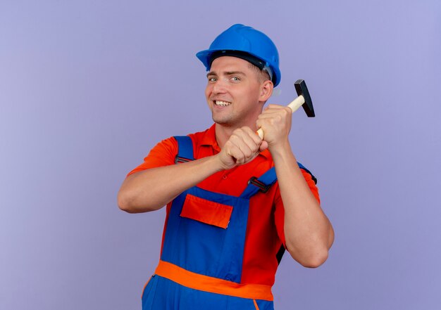 Smiling young male builder wearing uniform and safety helmet holding hammer around shoulder on purple