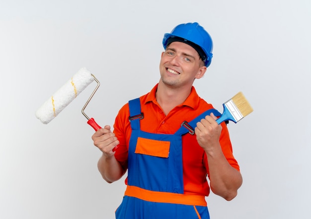 Smiling young male builder wearing uniform and safety helmet holding paint roller with paint brush on white
