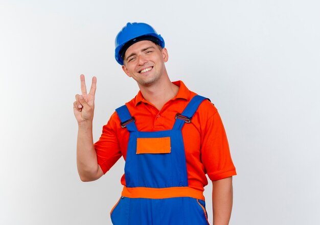 Smiling young male builder wearing uniform and safety helmet showing peace gesture on white